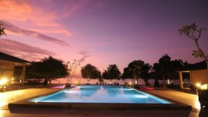 En swimmingpool i skumringen med udsigt til solnedgang, omgivet af træer og bygninger.