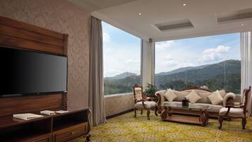 A spacious living room with a large flat-screen TV, a sofa set, and a coffee table. The room features a large window offering a view of mountains.