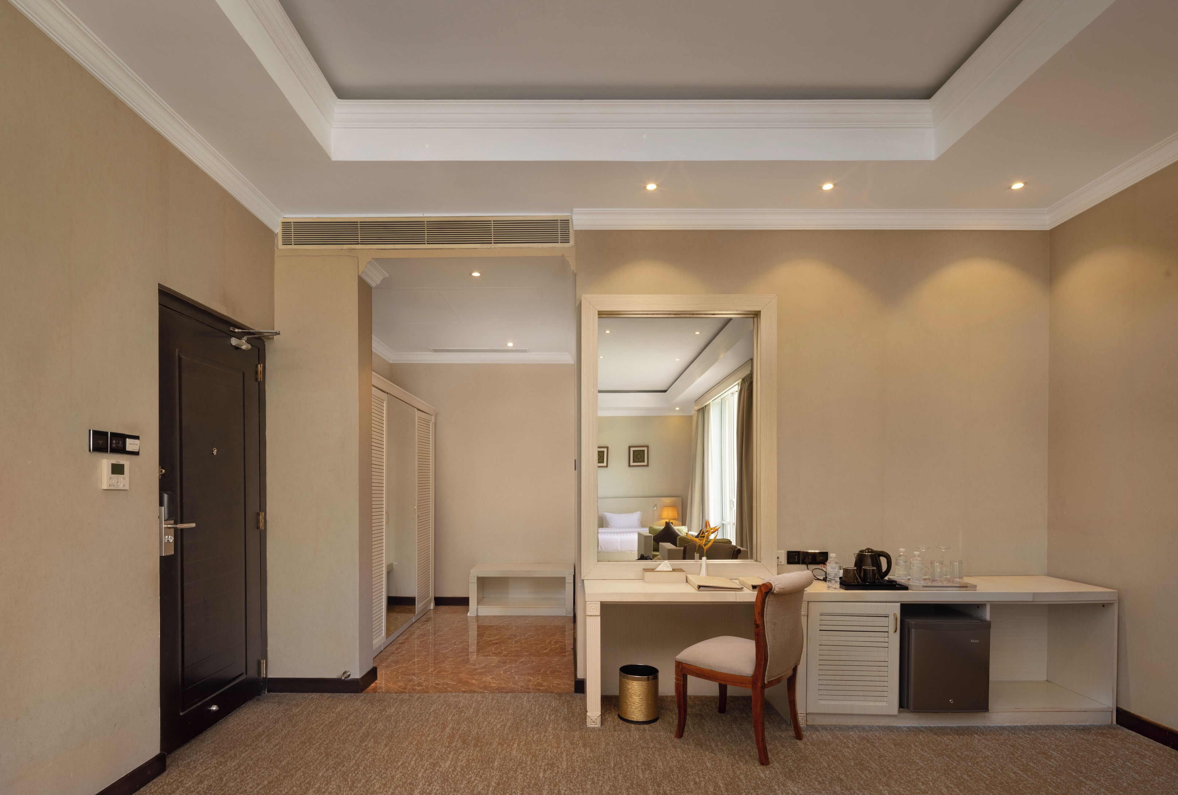 Une chambre d’hôtel équipée d’un bureau, d’une chaise et d’une petite kitchenette. Il y a une porte sur la gauche et un miroir au mur.
