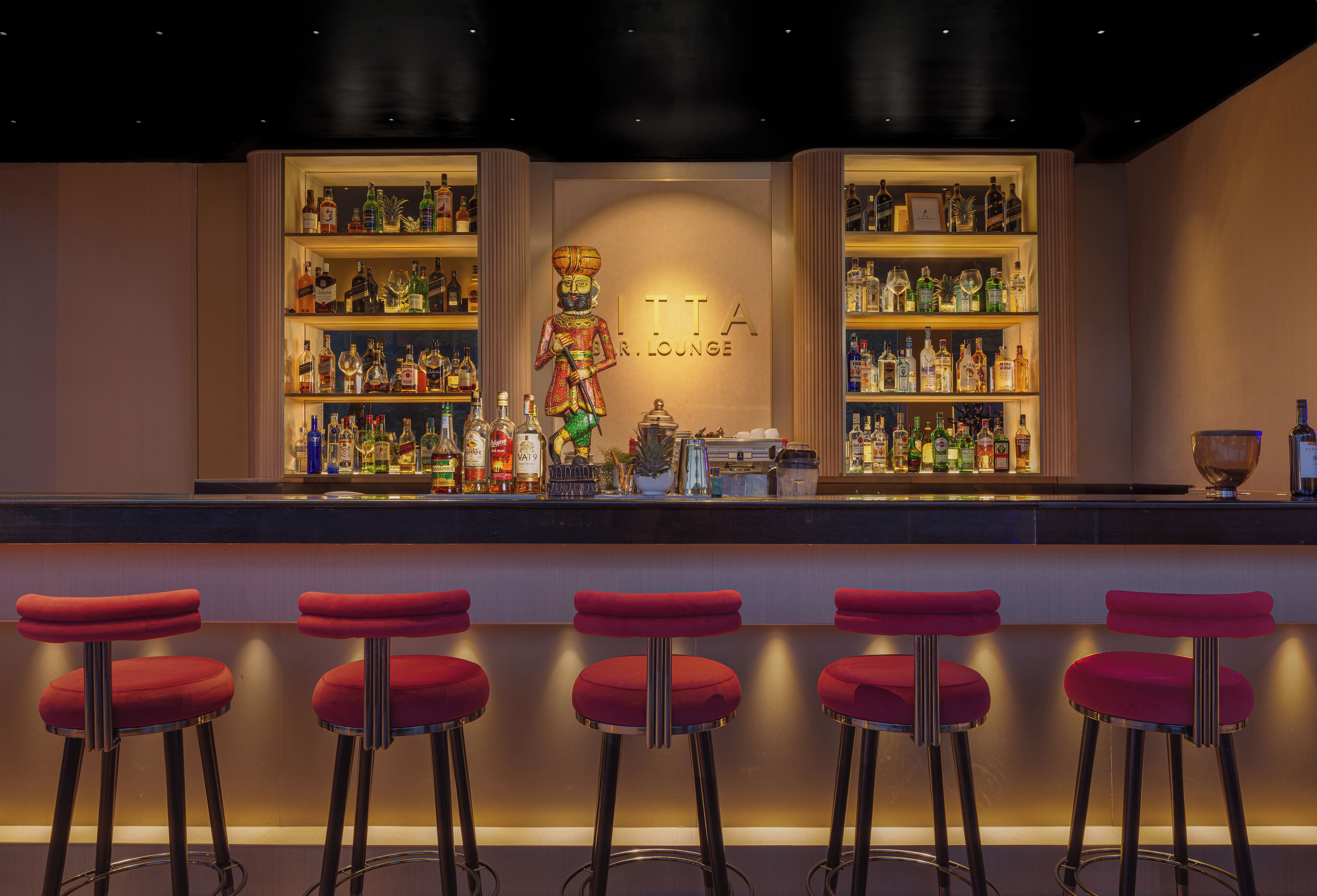 A bar area with a sign reading "LITA R. LOUNGE," shelves stocked with bottles, and five red bar stools.
