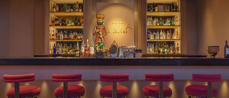 A bar area with a sign reading "LITA R. LOUNGE," shelves stocked with bottles, and five red bar stools.