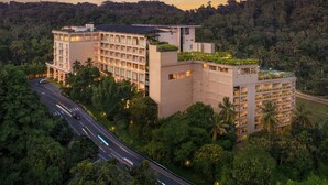Un hôtel à plusieurs étages doté d’un jardin sur le toit, entouré d’une végétation luxuriante et bordé par une route où circulent des véhicules.