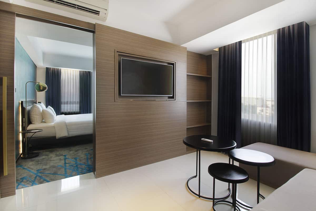 A modern hotel room with a bed, a TV mounted on the wall, a small seating area with two stools, and a large window with curtains.