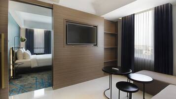 Una habitación de hotel moderna con una cama, un televisor montado en la pared, un pequeño rincón de estar con dos taburetes y un ventanal con cortinas.