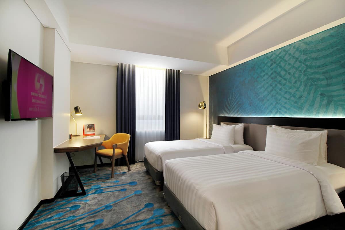 A hotel room with two beds, a desk, a chair, a TV, and a large window with curtains.