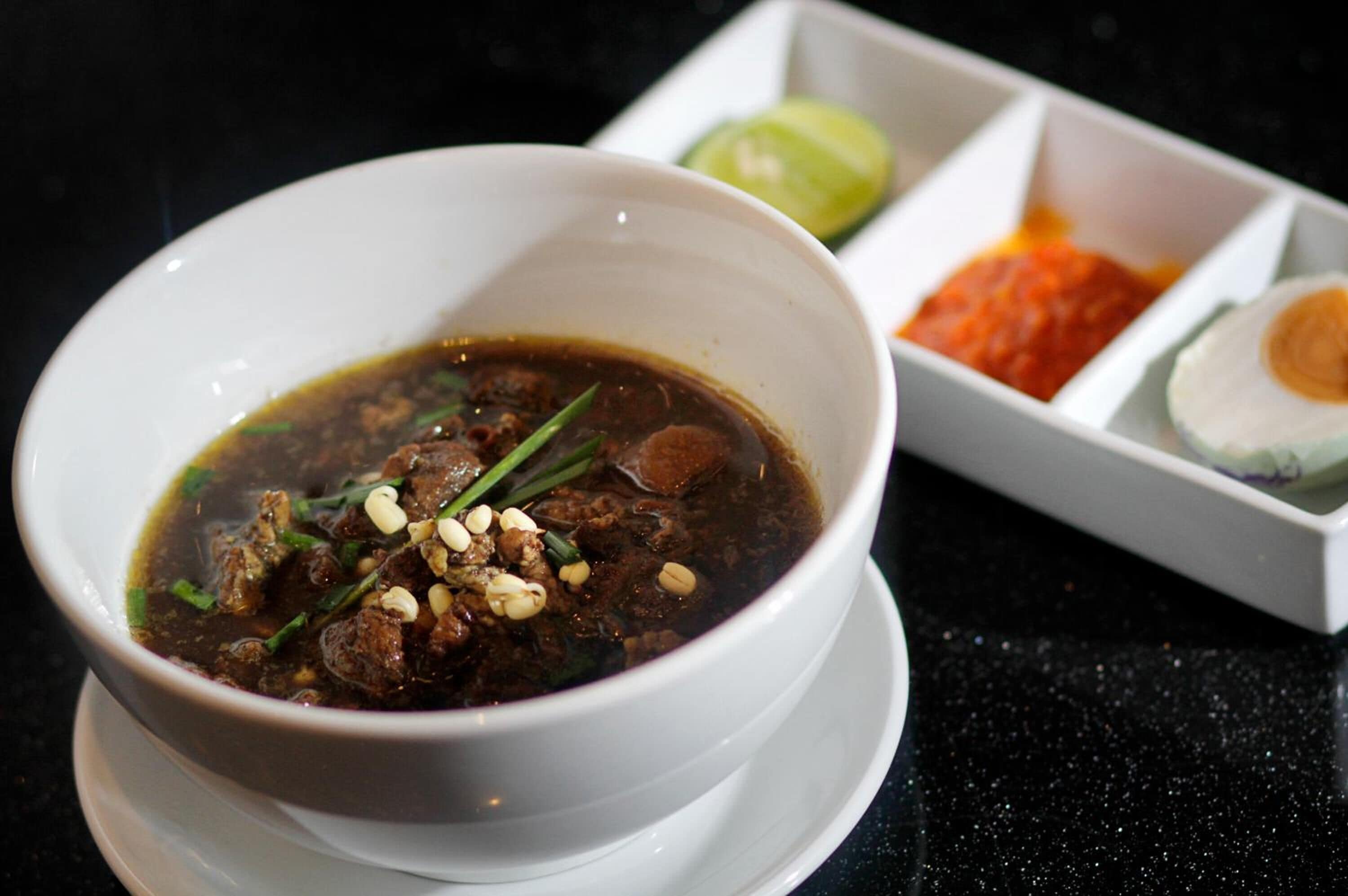 Un tazón de sopa con carne y guarniciones, acompañado de un limón y dos recipientes pequeños de salsas.