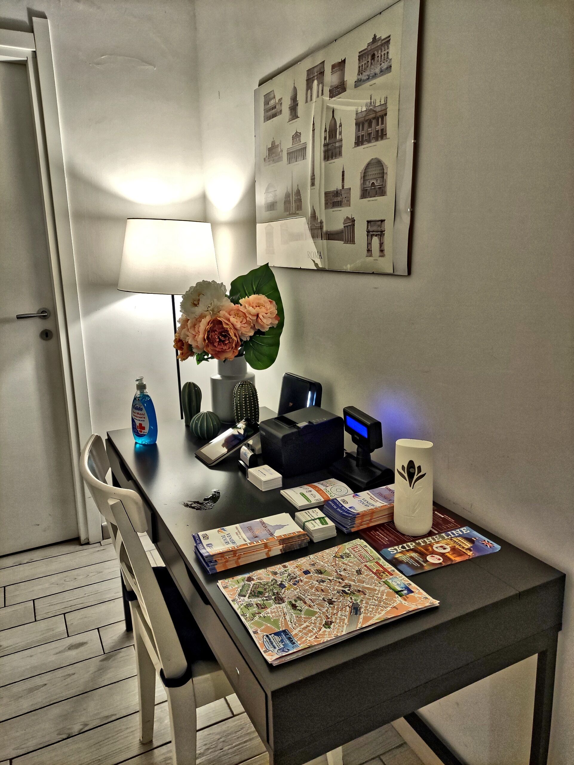 Habitación de hotel con escritorio, lámpara, florero con flores, mapa y un cartel en la pared.