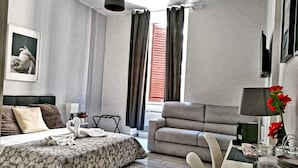 Quarto de hotel moderno com cama, sofá, área de refeições e janela com cortinas.