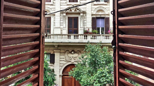 Visão através de venezianas de madeira de um edifício com várias janelas, uma varanda e uma grande porta de madeira.