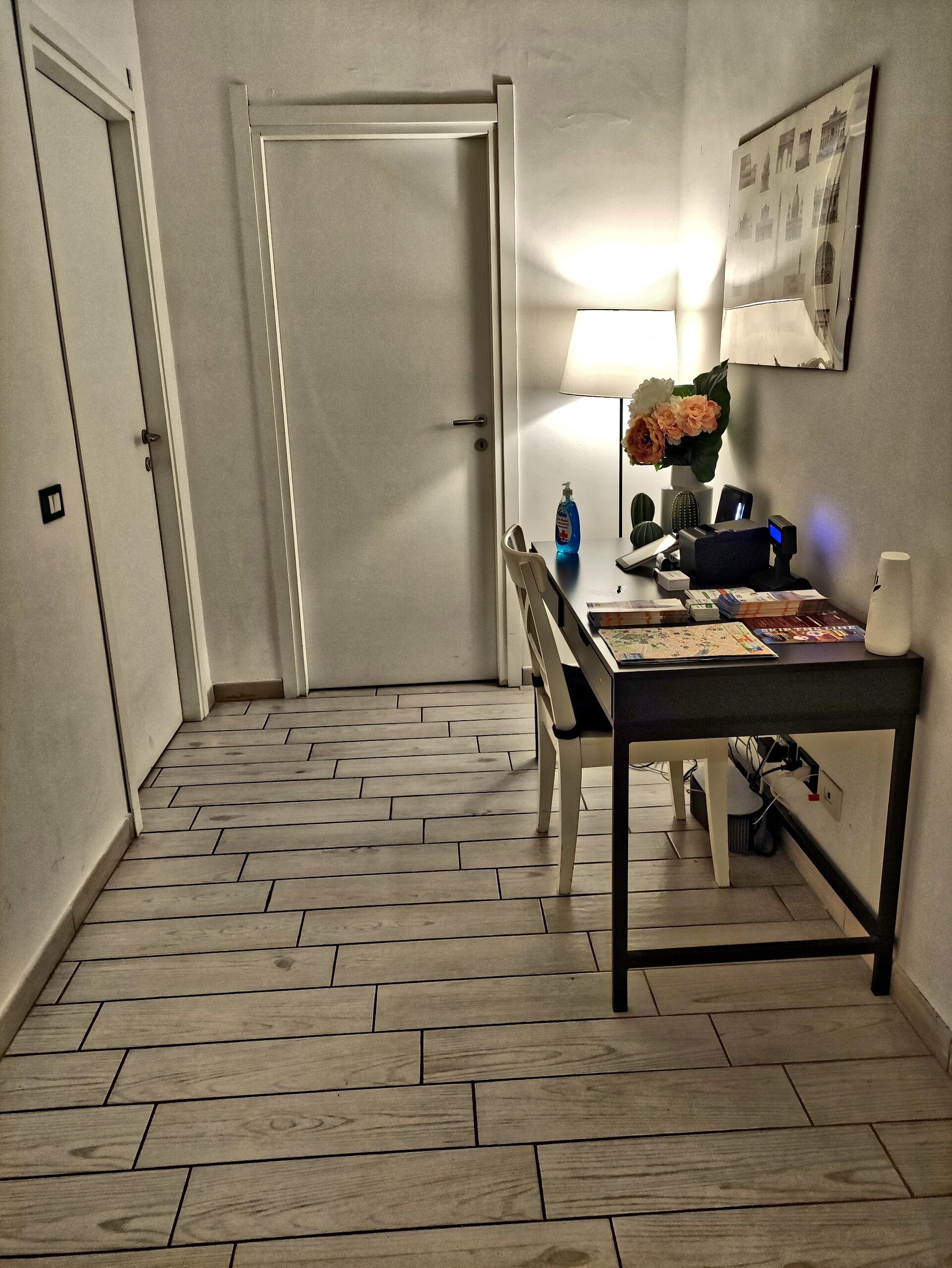 A hallway with a herringbone-patterned floor, a closed door, and a table with a lamp, flowers, and some items on it.