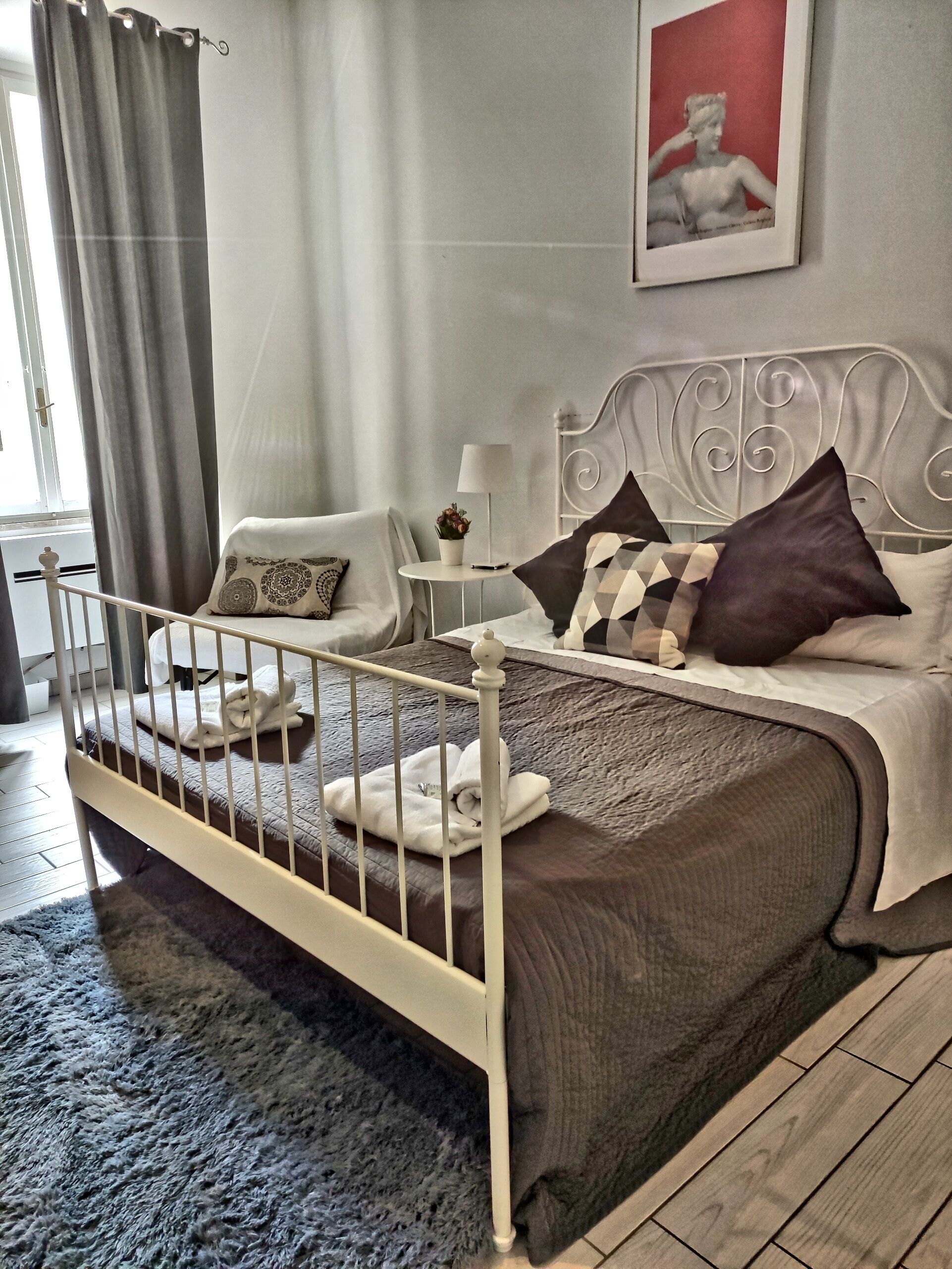 Un dormitorio con estructura de cama metálica, una mesita de noche con lámpara, una planta en maceta y un cuadro en la pared.