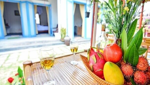 A table with a basket of fruits and two glasses of a yellow beverage, set outdoors with a building in the background. - Local Beach Homestay (Da Nang)