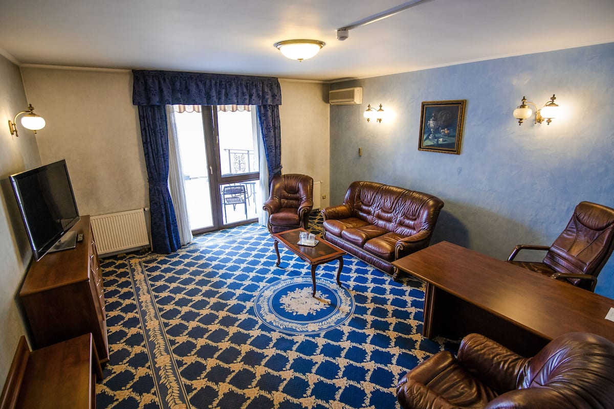 Una habitación con alfombra azul estampada, sofás de cuero, una mesa de centro de madera, un televisor y un amplio ventanal con cortinas.