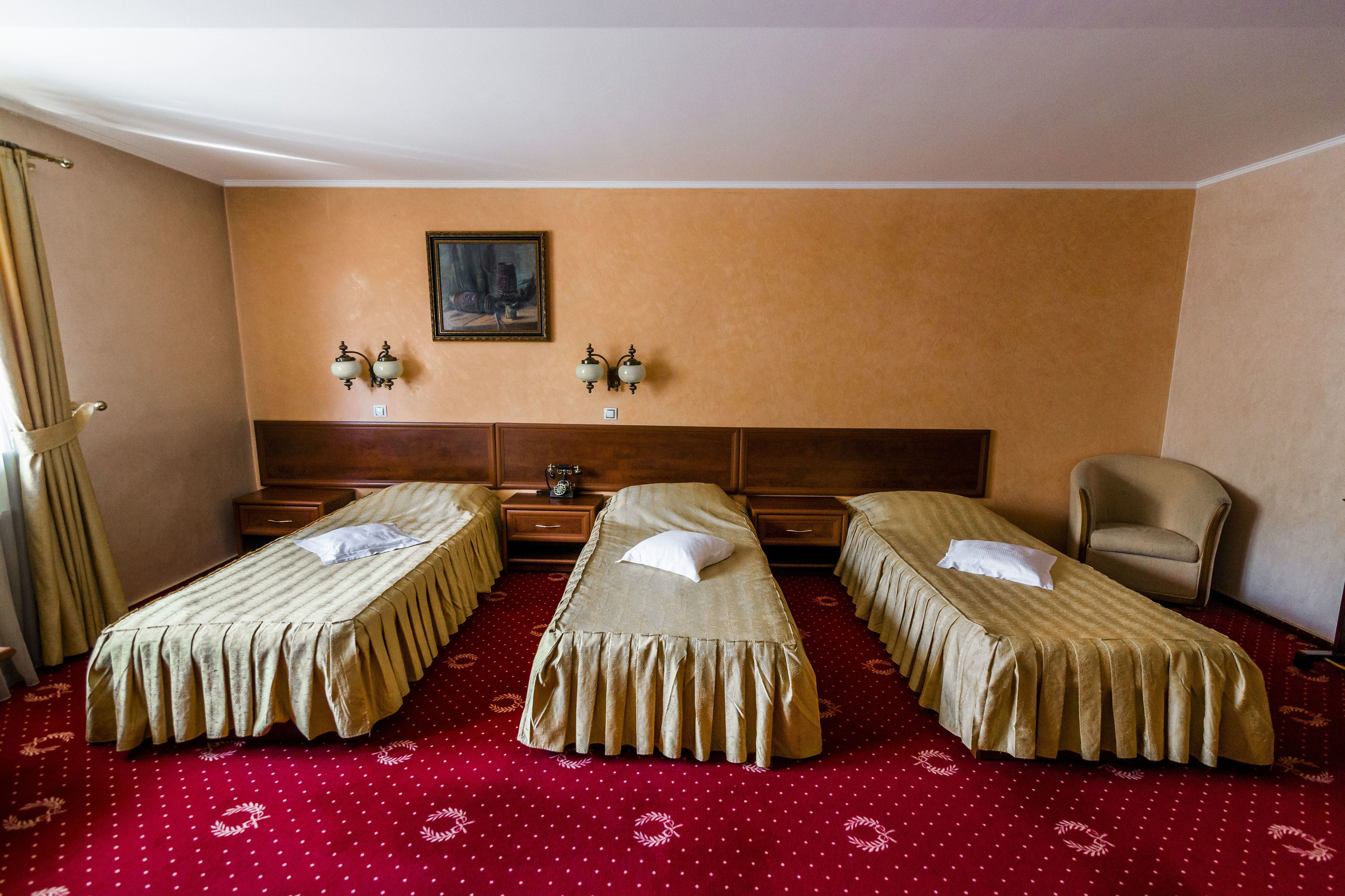 A hotel room with three single beds, a wooden headboard, a small armchair, and a painting on the wall.