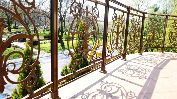 A wrought iron balcony railing with intricate designs, overlooking a well-maintained garden with trimmed hedges and trees. - Hotel Golden House (Craiova)