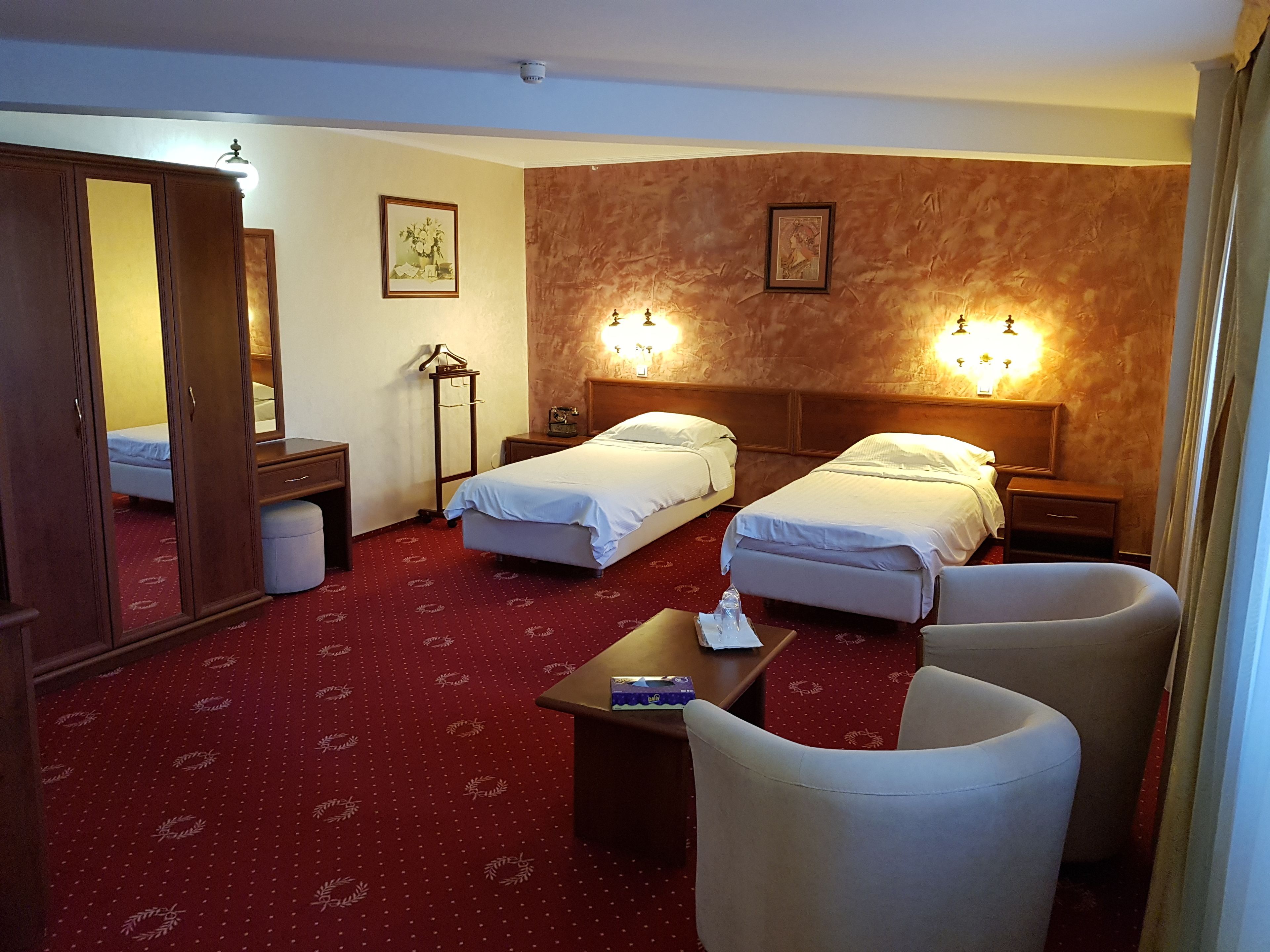 A hotel room with two beds, a desk, a chair, a mirror, a wardrobe, and a red carpet.