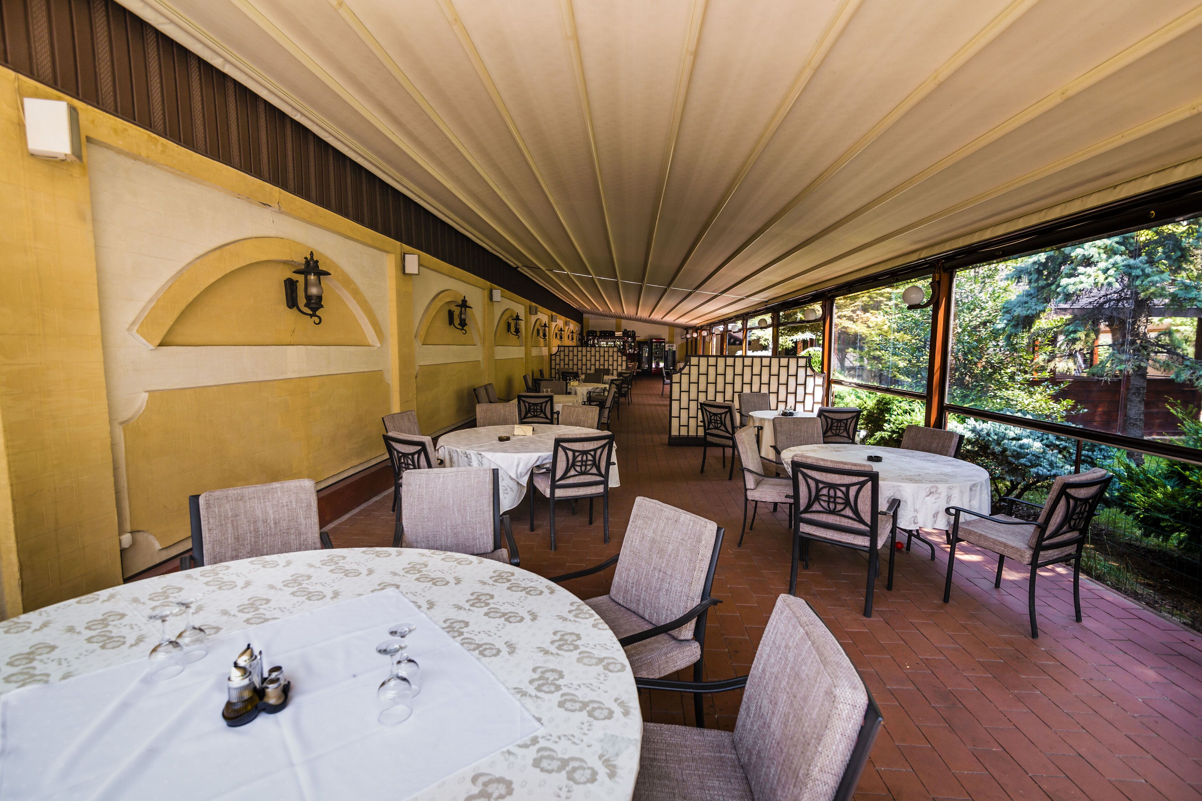 Un área de comedor al aire libre con mesas y sillas redondas, cubierta por un techo y rodeada de muros de ladrillo y follaje.