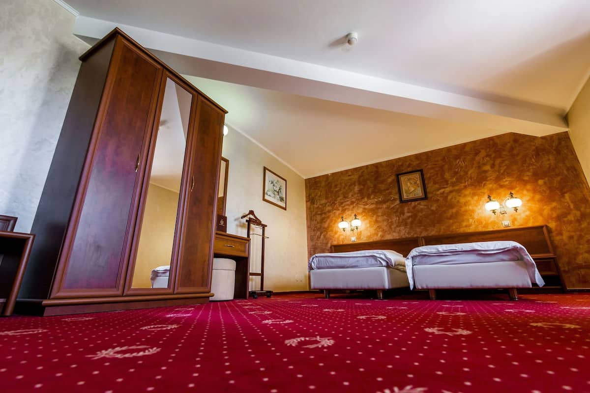 Habitación de hotel con un amplio armario de madera, dos camas, alfombra roja con estampado y luces fijadas a la pared.