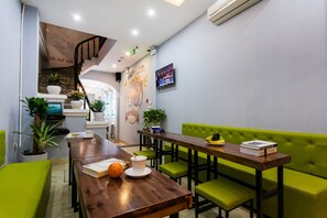 A modern dining area with a wooden table, green upholstered chairs, and a green sofa. There are potted plants, a television, and a staircase leading to another level. - Hanoi Centre Hostel (Hanoi)