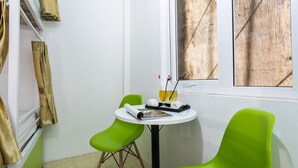 Una habitación pequeña con una mesa blanca, dos sillas verdes, un ventanal con persianas de madera y un perchero con una toalla.