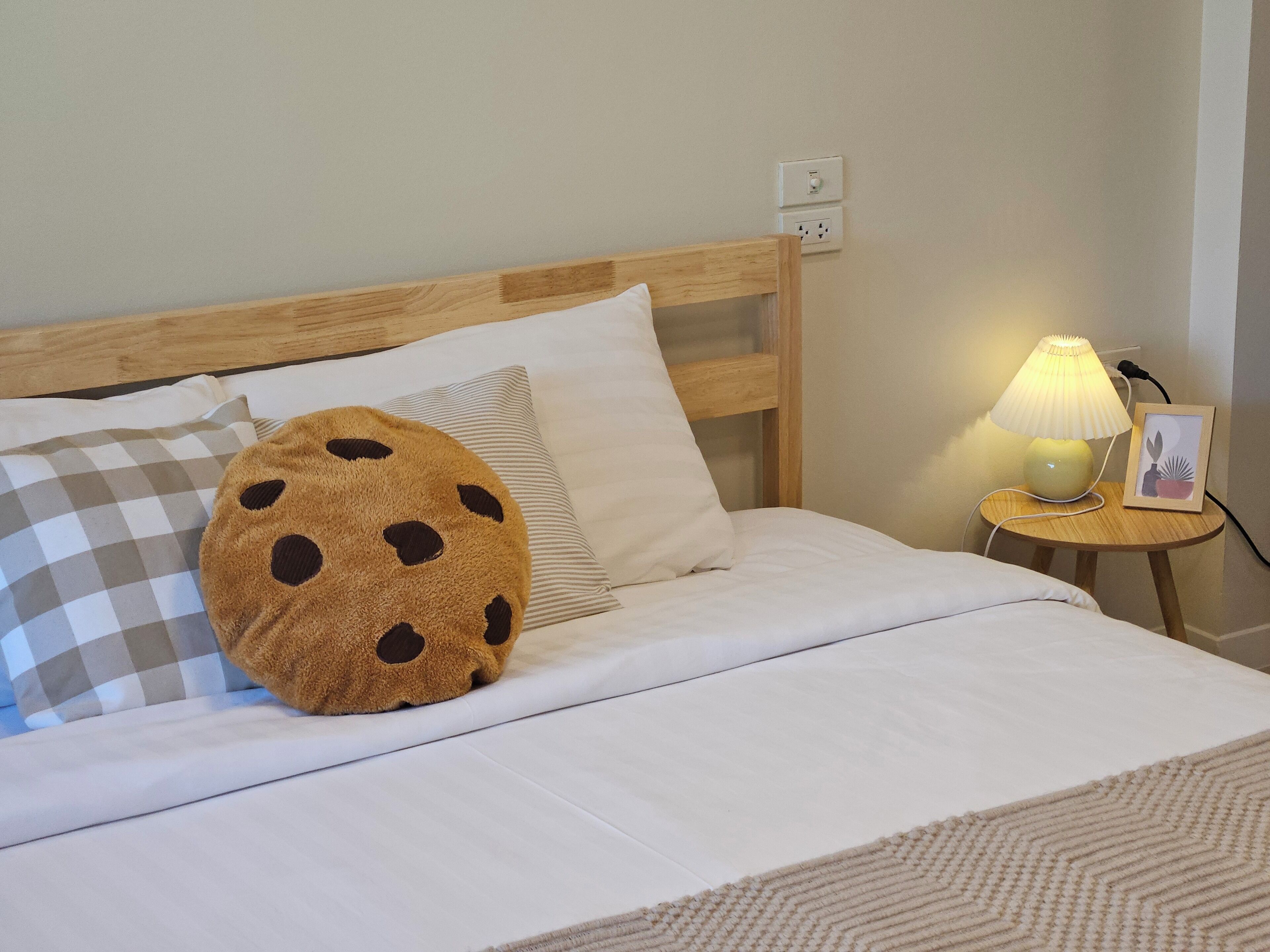 A neatly made bed with a wooden headboard, a round pillow with a cookie pattern, a striped pillow, and a small bedside table with a lamp and a picture frame.