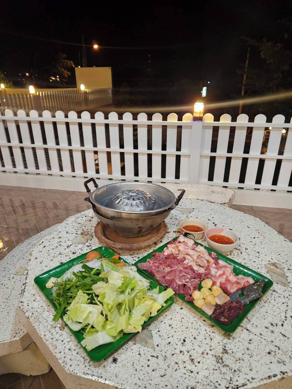 Mesa redonda com um aparelho de fondue, carnes e vegetais diversos em pratos verdes, cercada por uma cerca branca e postes de luz ao fundo.