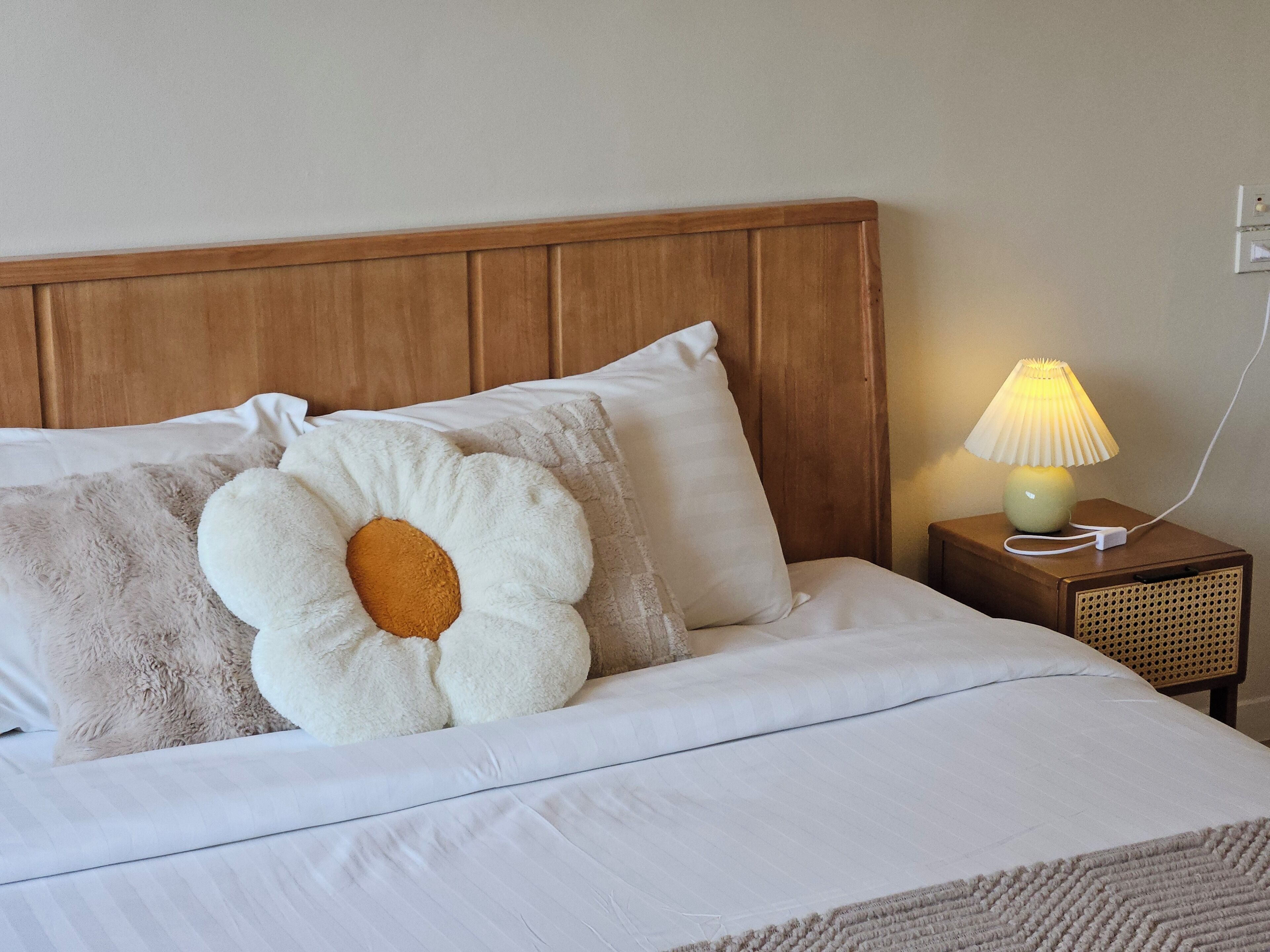Een net opgemaakt bed met witte beddengoed en een groot kussen in de vorm van een bloem. Er staat een nachtkastje met een lamp en een stopcontact.