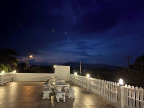 Una terrazza esterna con tavoli e sedie rotonde, un recinto bianco e vista sulle montagne sotto un cielo notturno.