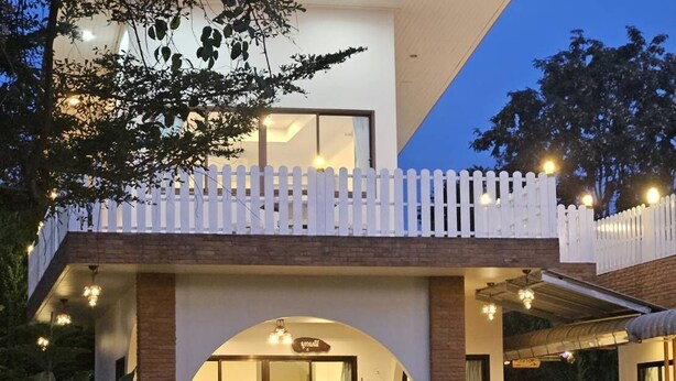 Une maison à deux étages avec une façade blanche, un balcon agrémenté d’une balustrade blanche et une terrasse couverte avec des sièges d’extérieur.