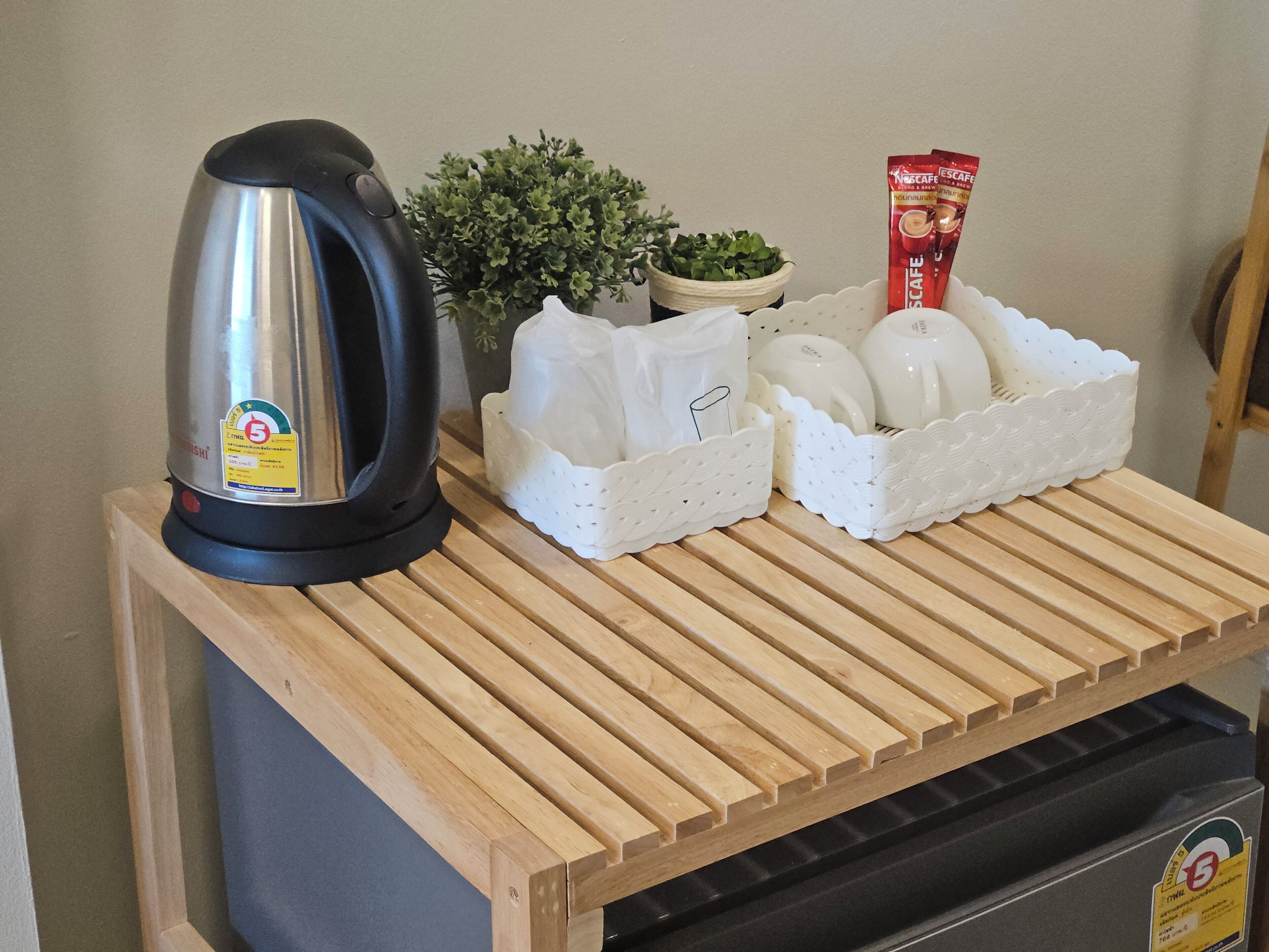 Una encimera de cocina con un hervidor, una pequeña planta en maceta, una cafetera y una cesta de tazas blancas.