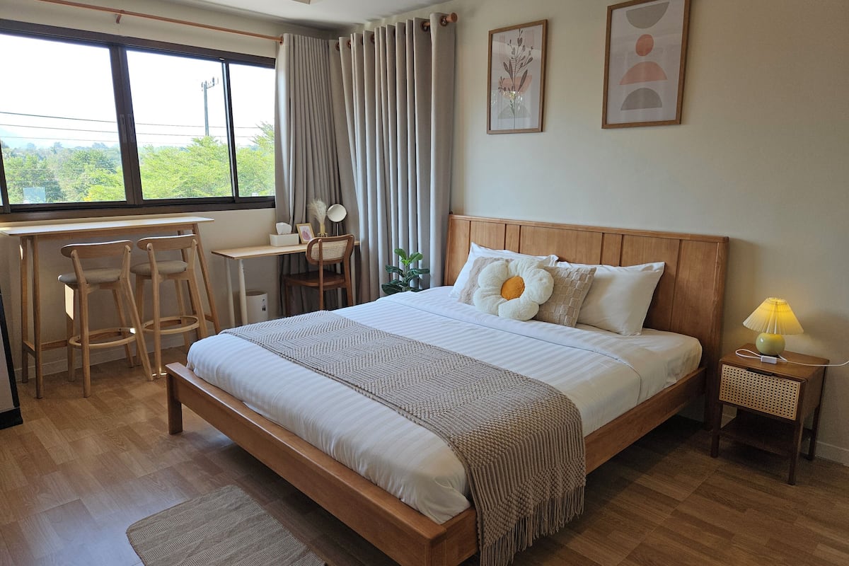 A bedroom with a wooden bed, a desk with two chairs, a bedside table with a lamp, and a window with curtains.