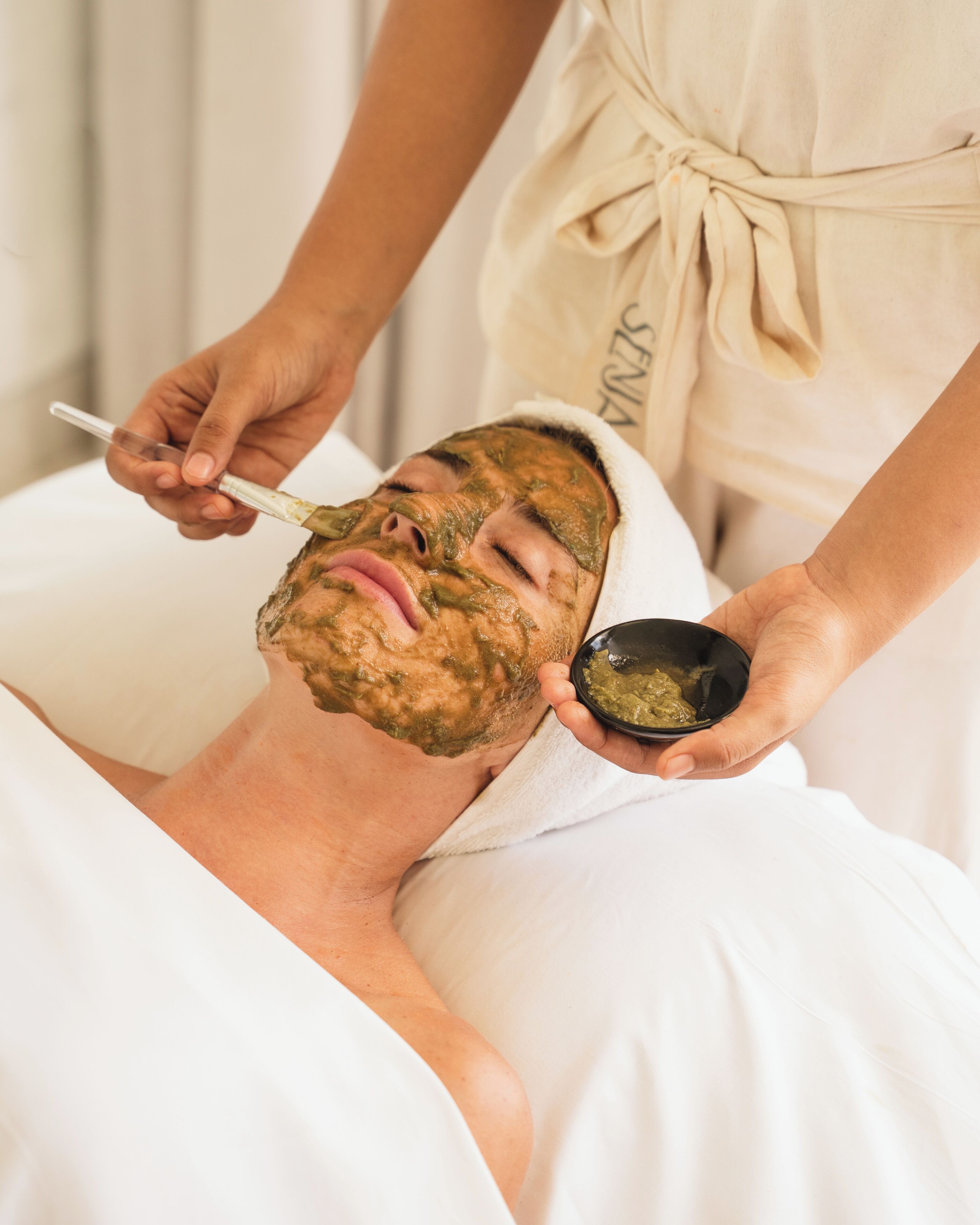 Una persona recibiendo un tratamiento facial con una mascarilla y un recipiente pequeño con pasta verde.