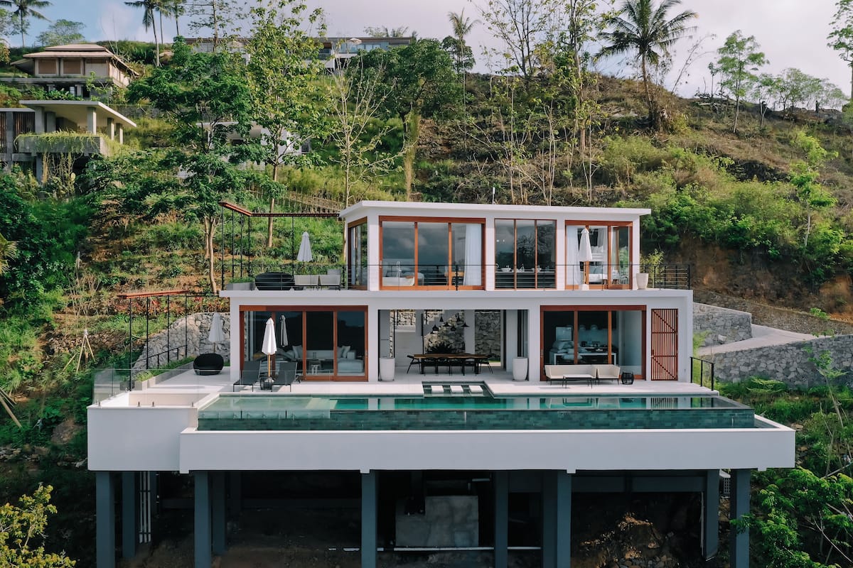 Una casa moderna de dos pisos con piscina en la azotea, rodeada de exuberante vegetación y colinas.