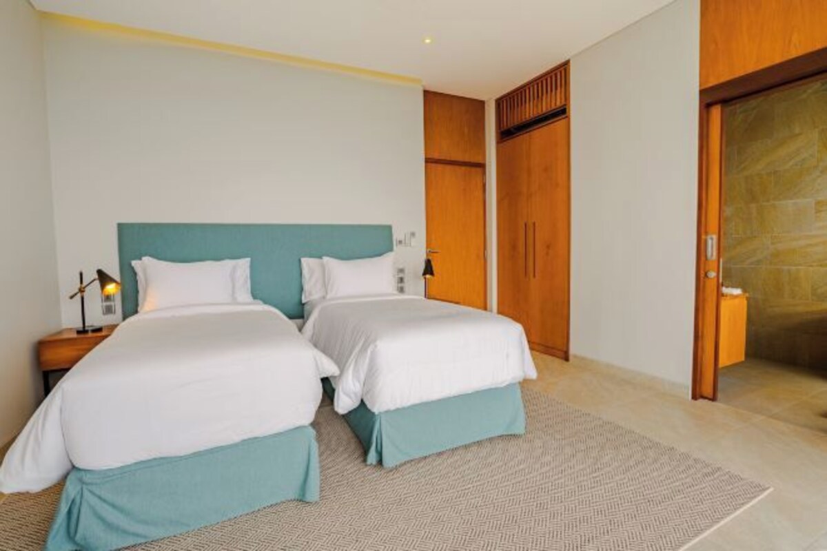 Habitación de hotel con dos camas, un armario de madera, una mesita de noche con lámpara y una puerta que da a otra habitación.