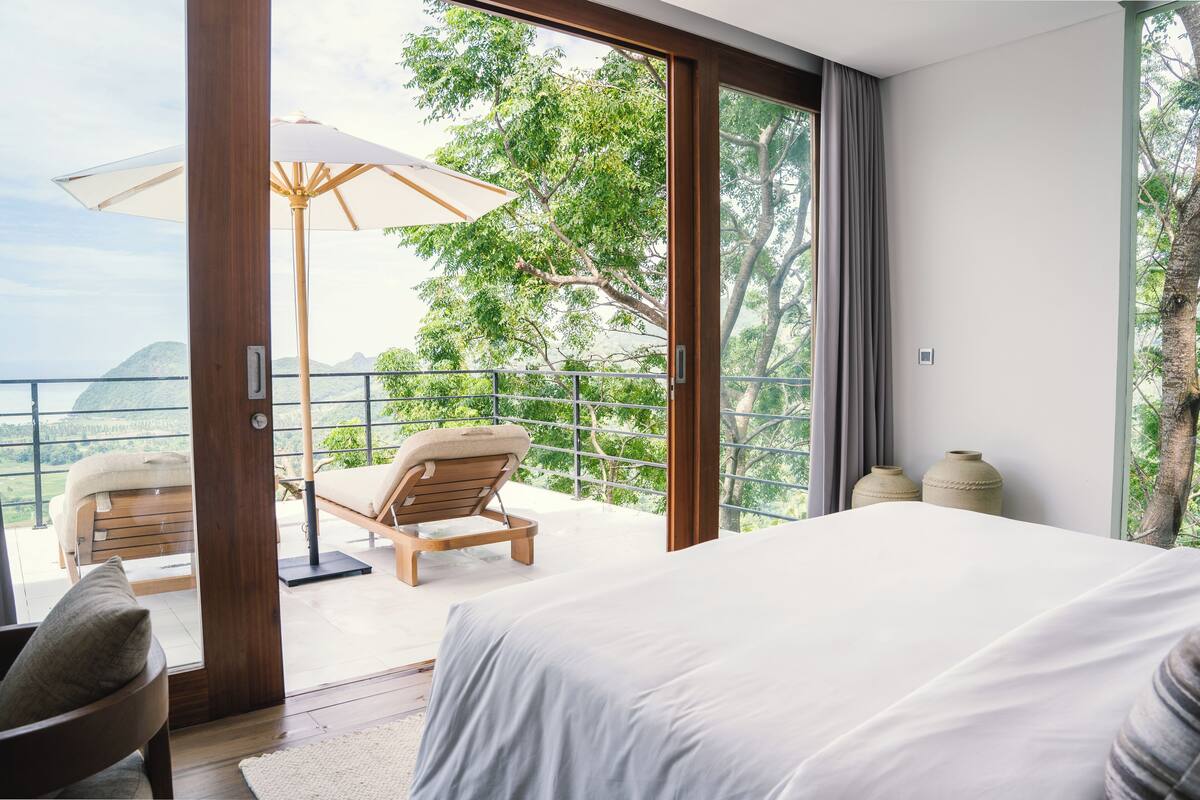 Un dormitorio con una cama grande, una silla de madera, una mesita y un balcón con vistas a árboles y montañas.