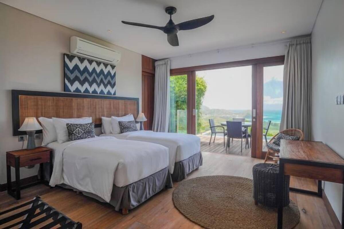 Habitación de hotel con dos camas, un ventilador de techo, equipo de aire acondicionado, un ventanal con vistas y un rincón comedor con mesa y sillas.