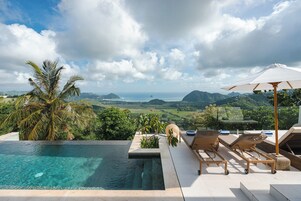 Piscina infinita com vista para o oceano e montanhas, cadeiras de praia e um guarda-sol branco.