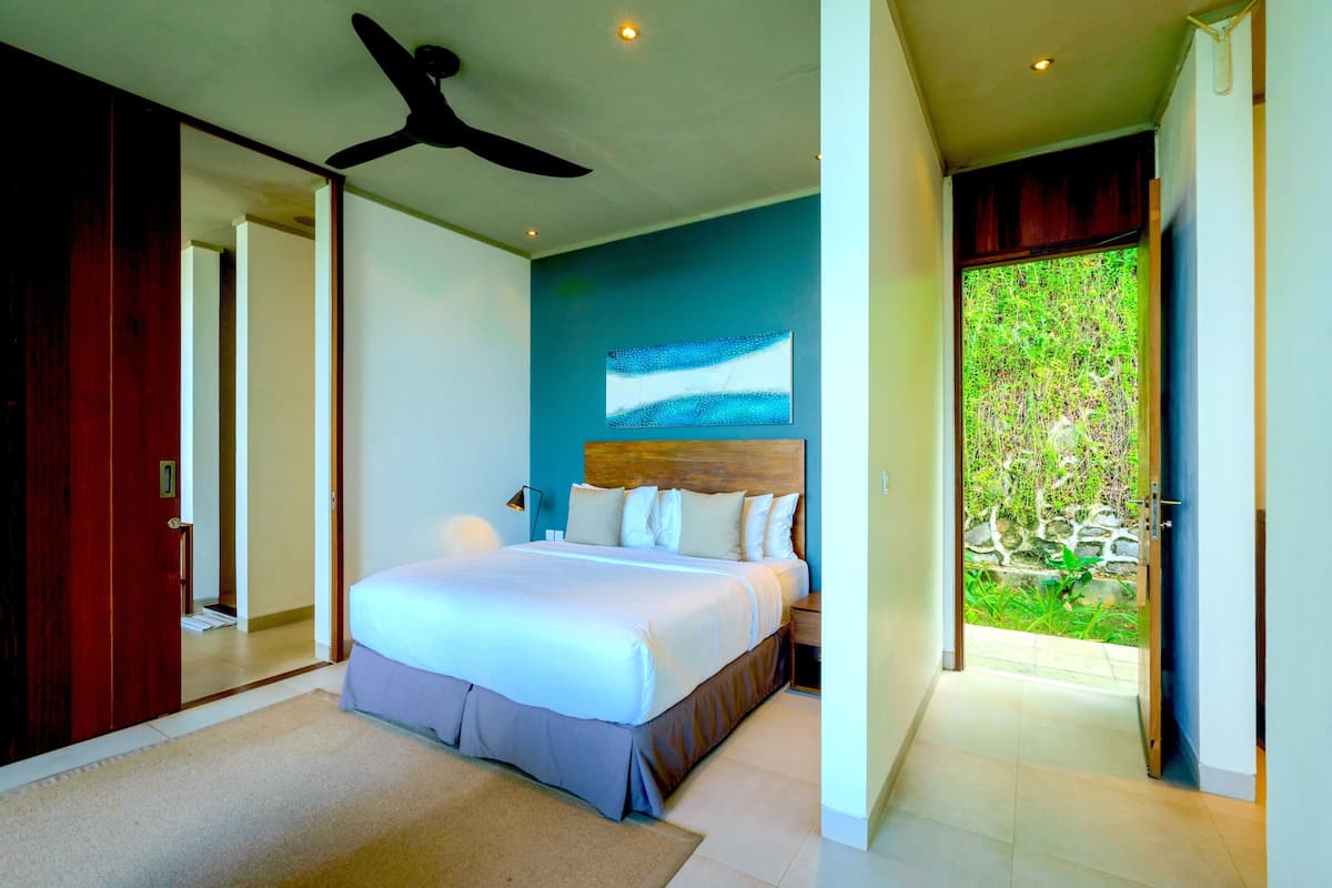 Un dormitorio con una cama grande, un ventilador de techo y vista a un jardín por una puerta abierta.