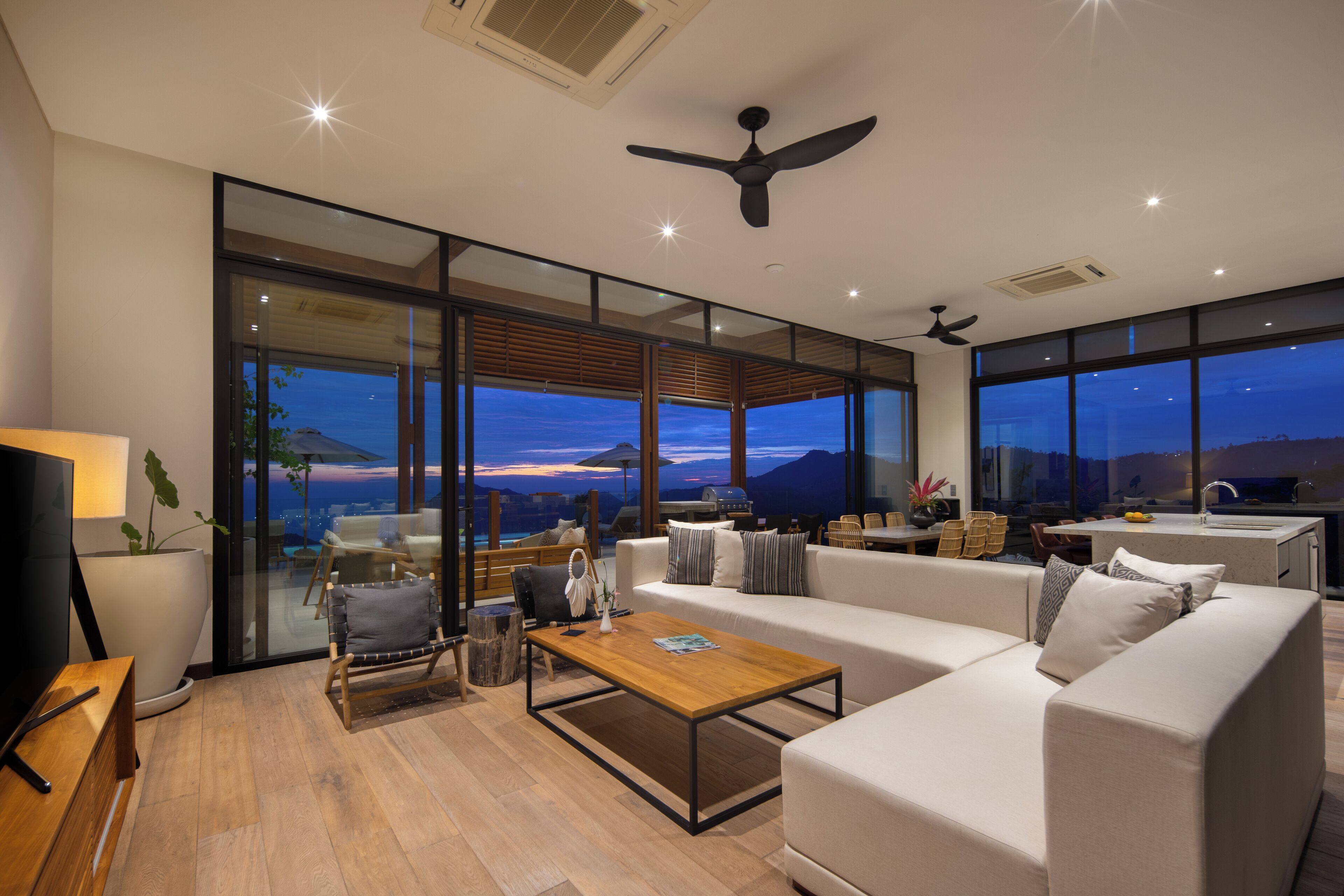 A modern living room with a sofa, coffee table, and a TV. There are large windows with a view of the outdoors.