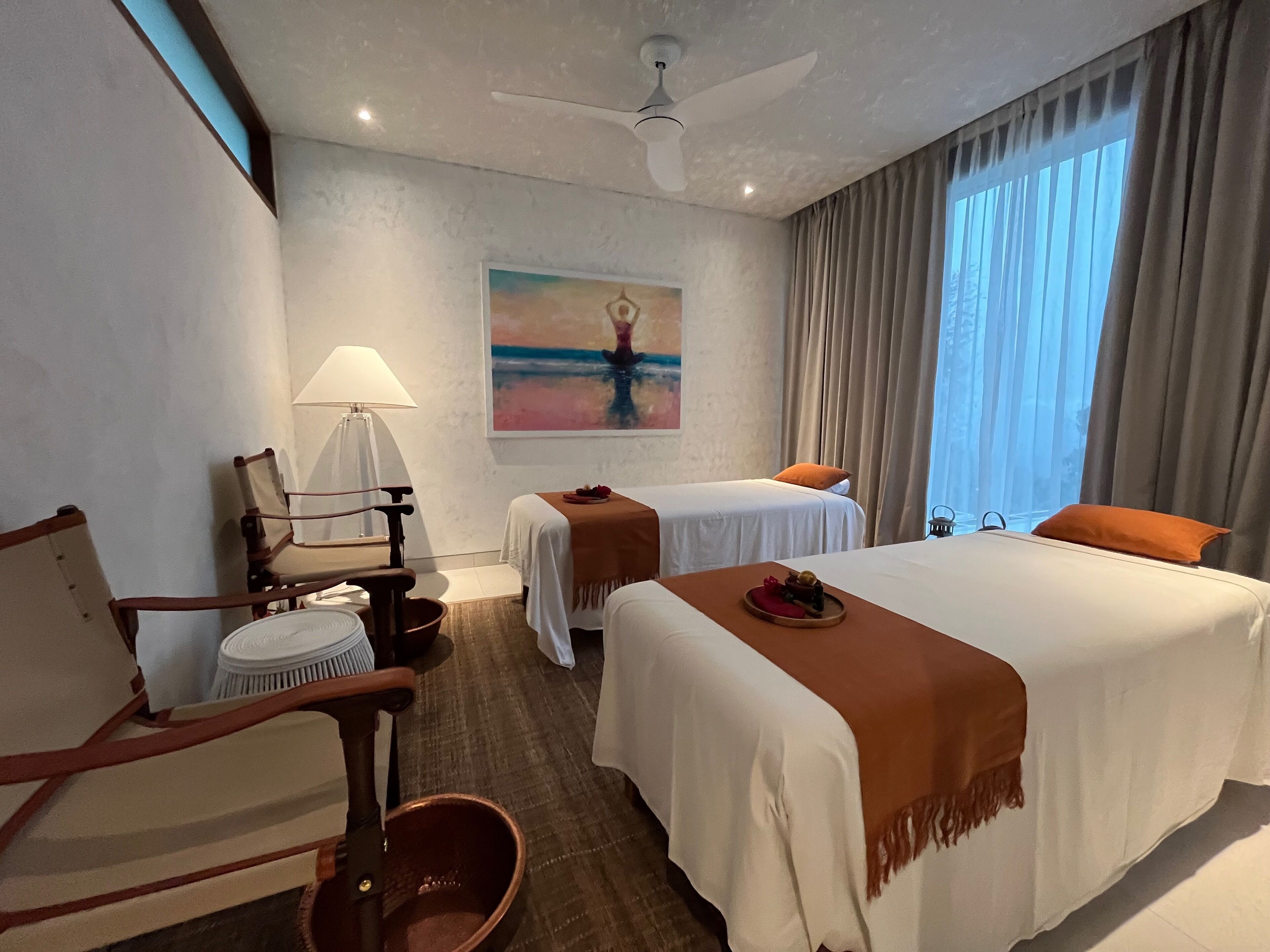 Quarto de hotel com duas camas, uma cadeira, uma mesa, um abajur e um quadro na parede.