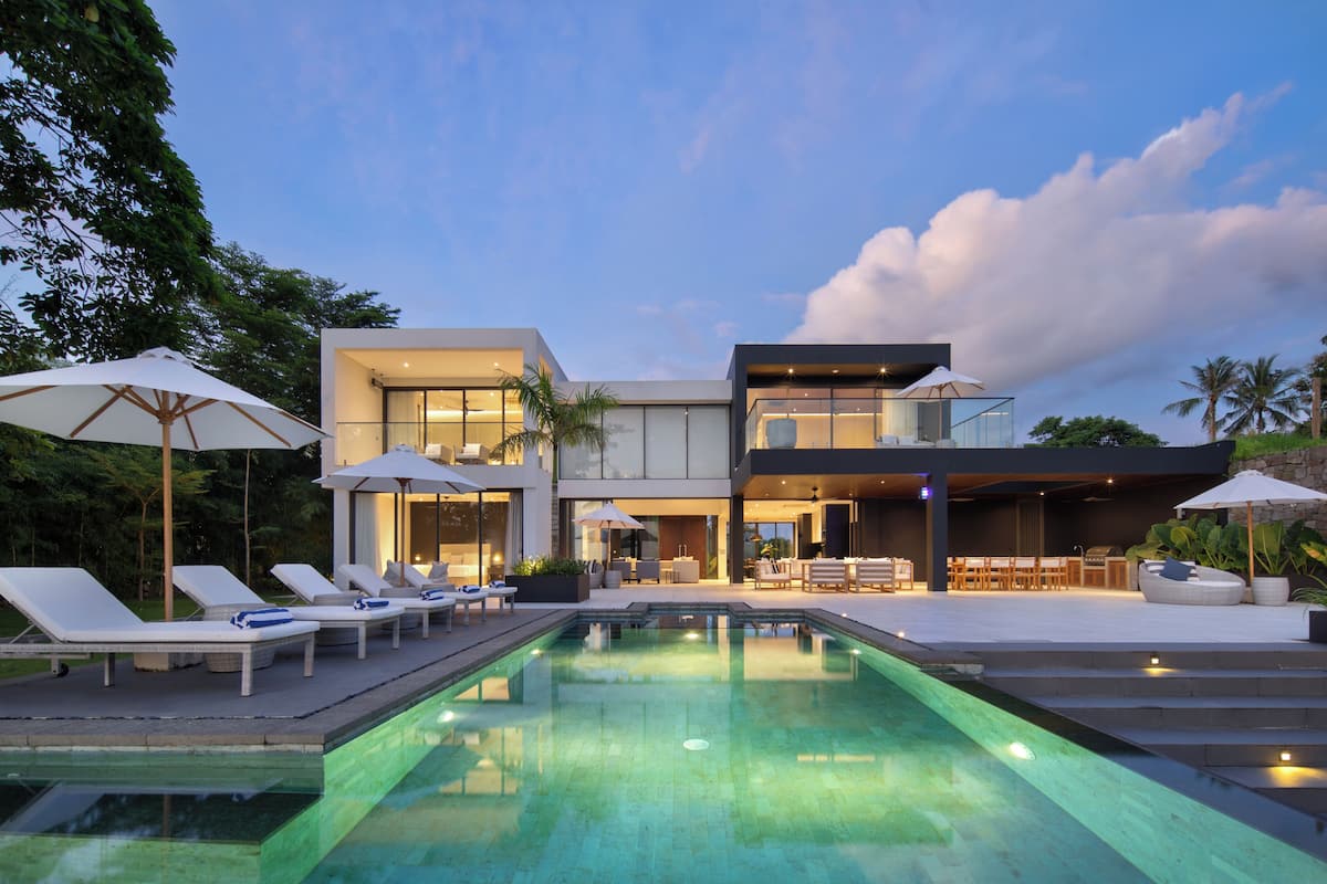 Una casa moderna de dos pisos con piscina, mobiliario exterior y sombrillas.