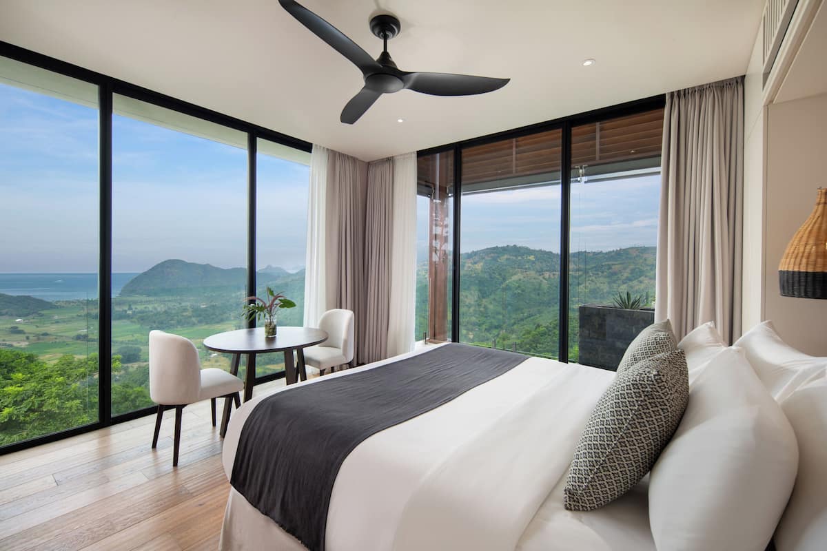 Un dormitorio con una cama grande, un ventilador de techo, una mesa de comedor con sillas y vistas a un paisaje montañoso a través de ventanas amplias.