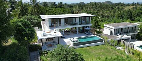Una casa moderna de dos pisos con piscina, rodeada de exuberante vegetación y colinas.