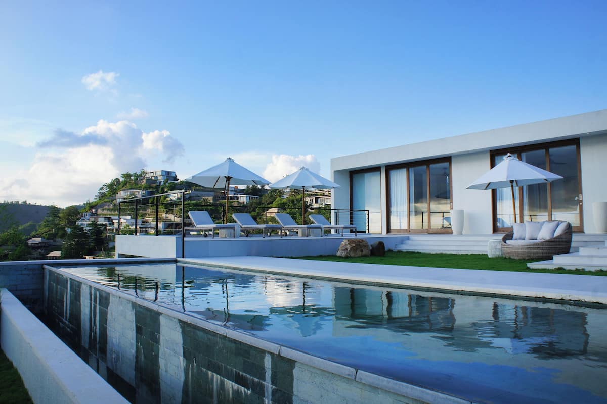 Un área de piscina moderna con sillones reclinables y sombrillas, rodeada por un césped bien cuidado y un edificio contemporáneo con grandes ventanales.
