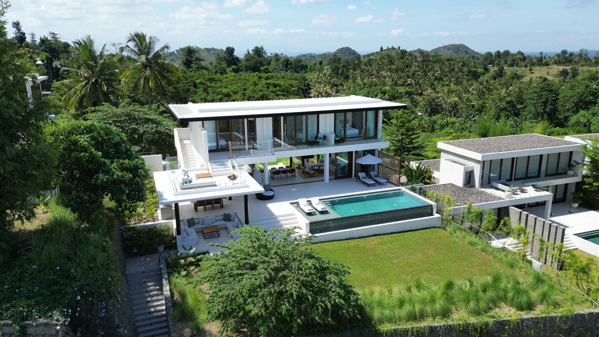 A modern two-story house with a large swimming pool, outdoor seating, and lush greenery surrounding the property.