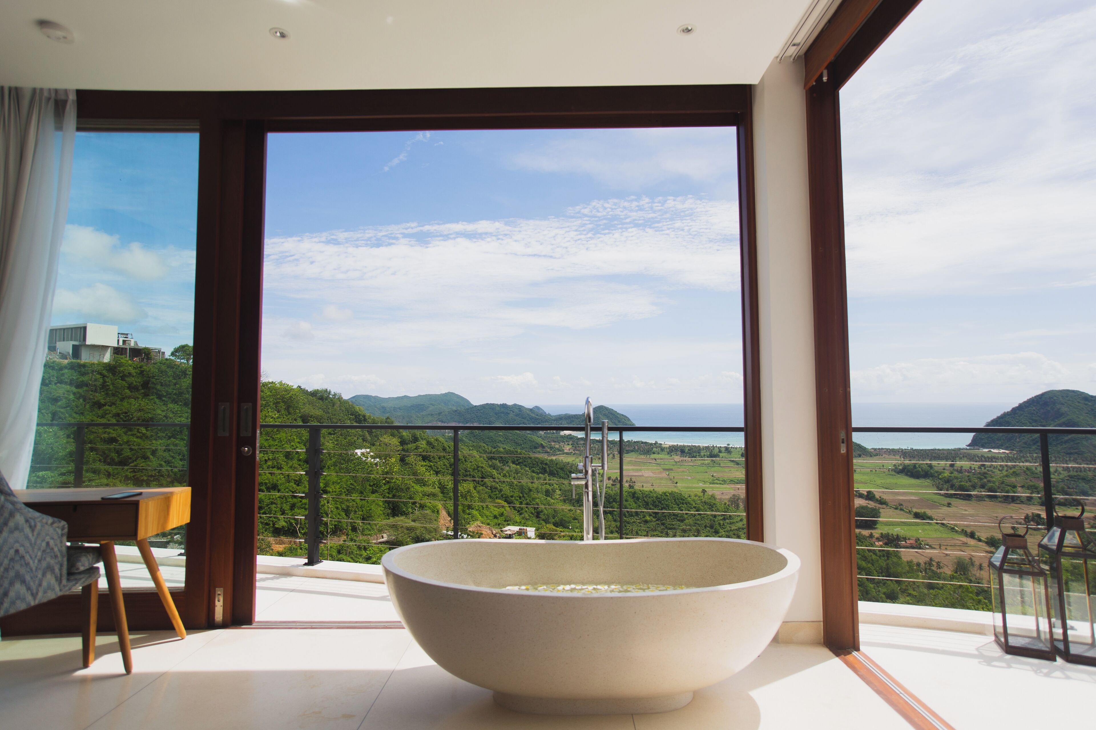A modern bathroom with a large bathtub, a wooden chair, and a table. It features a large window with a view of a green landscape and mountains.