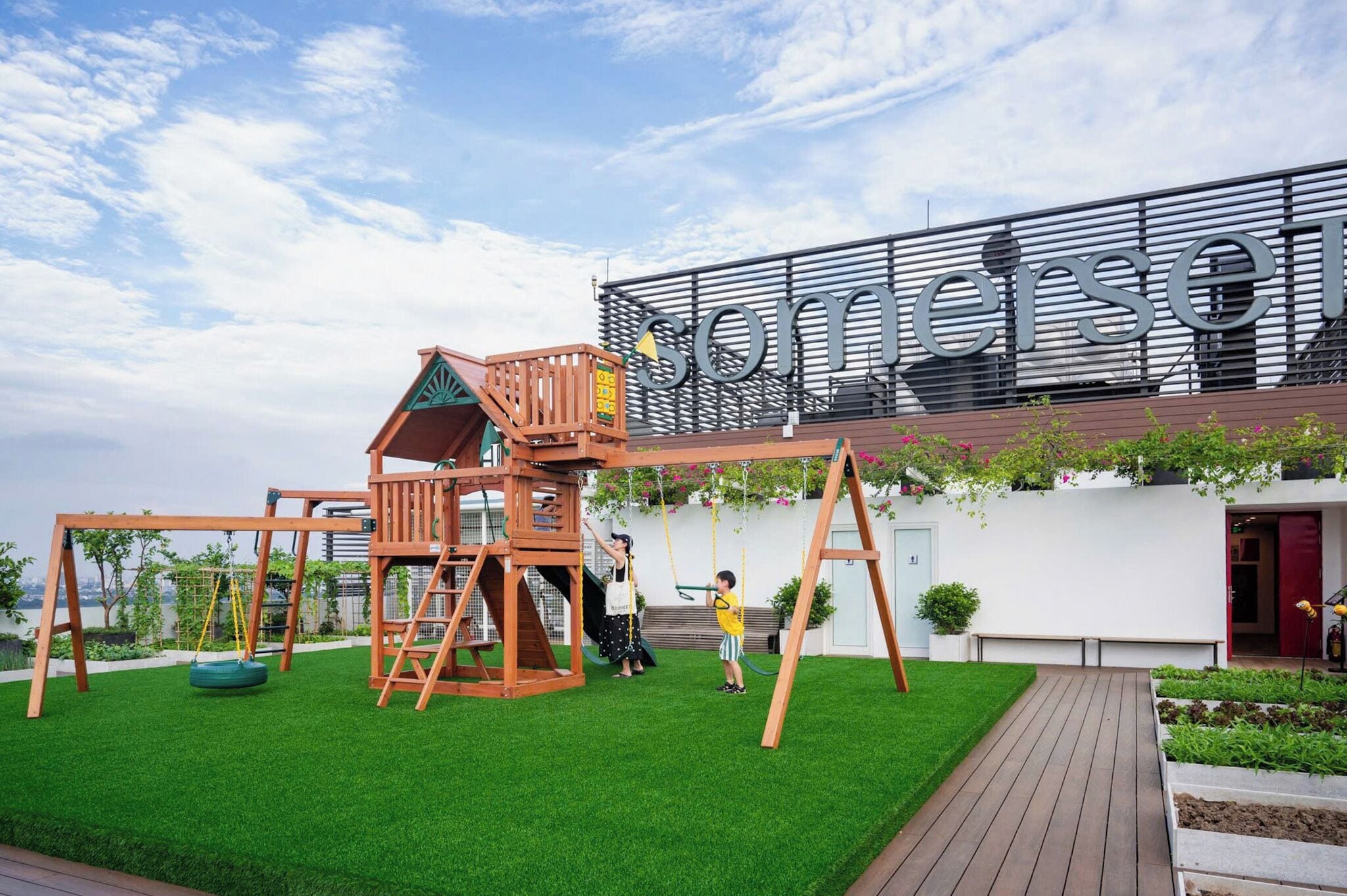 A playground with a wooden play structure and swings, a white building with 'Somersein' signage, and a clear sky.