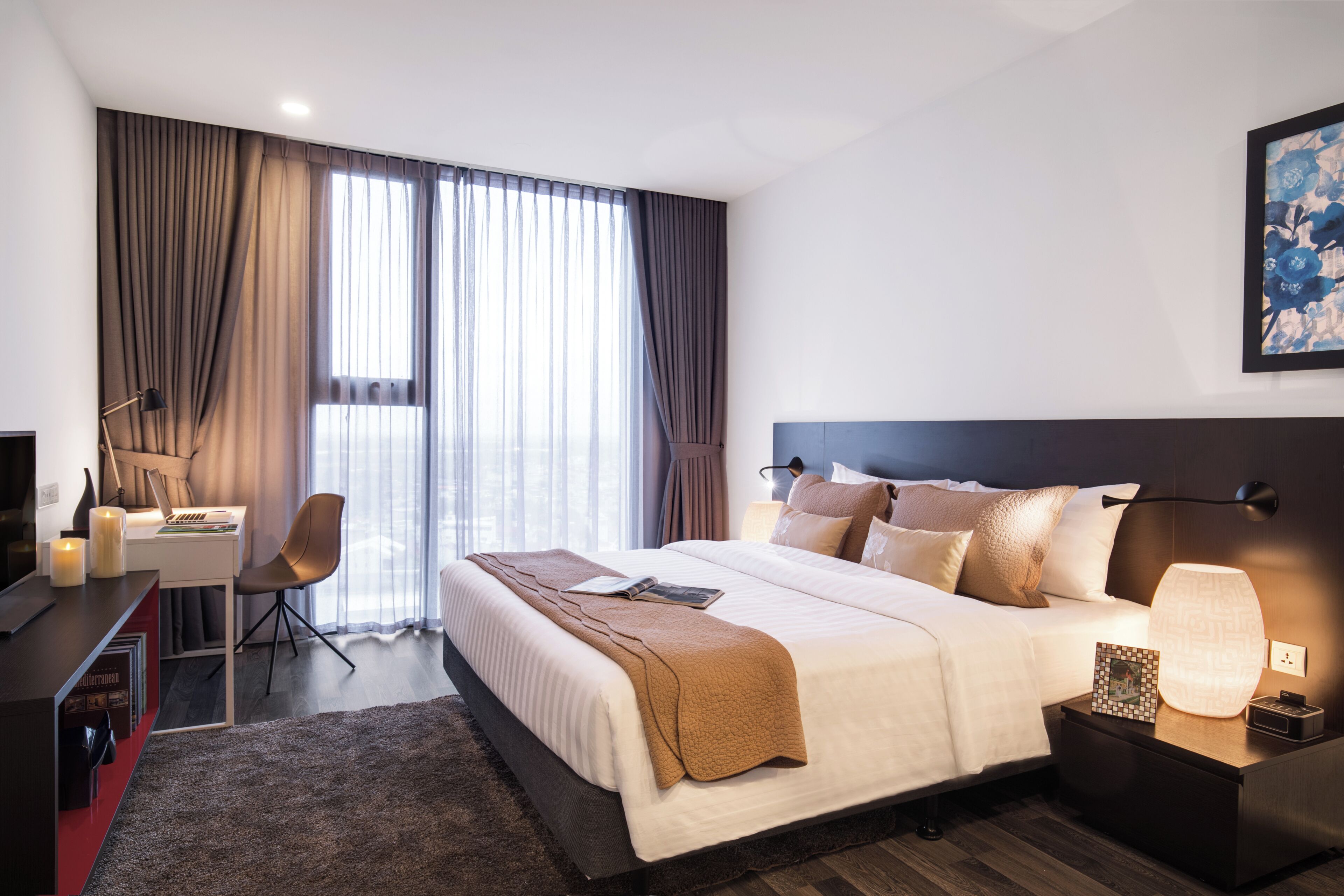 A modern hotel room with a large bed, a desk with a chair, a desk lamp, a bedside table with a lamp, and a large window with curtains.