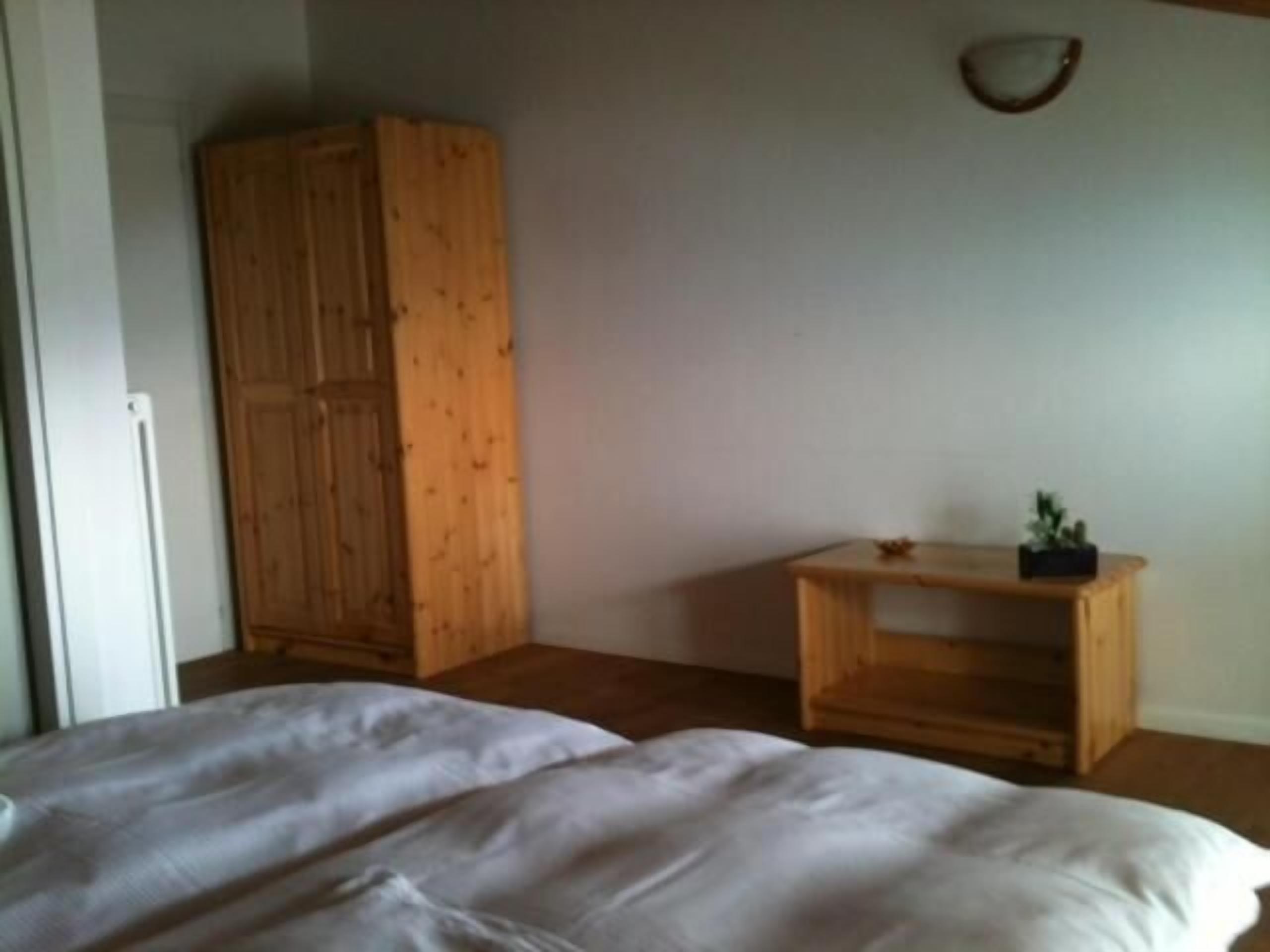 A bedroom with a wooden wardrobe, a bedside table with a plant, and a bed with white bedding.