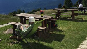 A person in a green chair reading a book at a wooden picnic table with a mountain view in the background. - Malga Zanga (Arco)